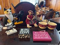 Two cast iron cat door holders, a fishwife doll holding wine corks, a stuffed doll with a chalkboard sign, two wooden block signs, and a country woven wastebasket in background.