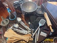 Photo showing an assortment of kitchen utensils on round wooden table including wooden spatulas, ladles, metal steamer basket, mortar and pestle, rolling pin, and plastic spatulas.