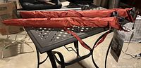 Photo showing two folded orange-red umbrellas in protective cases on a metal patio table.
