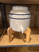 Side view of white ceramic water crock with two blue stripes, plastic spigot, on wooden stand.