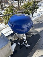 Blue Weber kettle grill with lid closed and black protective grilling mat under it on outdoor deck with snow on ground.