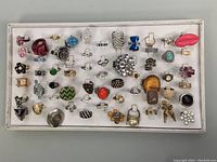 Tray showing over 50 assorted costume rings of various styles and colors arranged in multiple rows on white background.