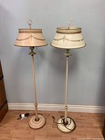 Full view of the pair of tall toleware metal floor lamps, one in blush and one in ivory, showing bases, poles and lampshades with gold decorative detailing.
