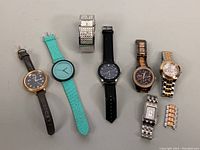 Seven assorted watches arranged horizontally on a surface showing a mix of strap materials and colors, some metal bands, one with crystal decoration.