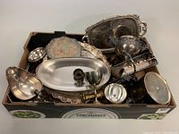 Box filled with assorted silver plated servingware including goblets, trays, a gravy boat, and other pieces showing tarnishing and varied sizes.