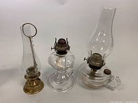 Photo showing the brass candleholder with attached snuffer and two kerosene oil hurricane lamps with glass chimneys placed side by side on a white background.