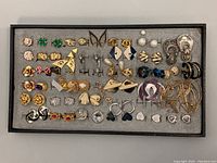 Full collection of assorted vintage costume earrings laid out on a grey foam board inside a black tray showing varied styles and colors including faux pearls, gold-tone, rhinestones, and clip-ons.