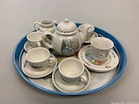 Full set displayed on round blue tin tray showing cups, saucers, creamers, and teapot with story illustrations and text