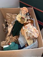 Box of holiday figurines including two caroler figures, one holding pinecone and berries, and chef/baker figurine holding pot, surrounded by holiday decorations and a miniature tree.