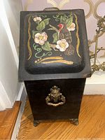 Front view of black enamelware coal scuttle with painted floral and bird motif on top, showing lion head handles and lion paw feet.