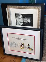 Two framed artworks leaning against a wall: one Disney Mickey Mouse animation art and one black-and-white portrait photo in a decorated frame.