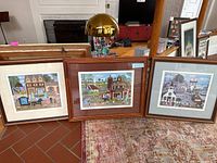 Three framed prints showing colorful village scenes by Cate Mandigo and a Grant Wood print, displayed side-by-side.