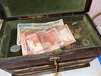 Open wooden jewelry box displaying assorted foreign banknotes and coins on green felt lining.