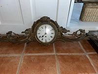 Full front view of ornate mantle clock on tiled floor showing scrollwork and clock face