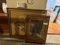 Two framed artworks placed side by side, the larger floral print behind, the smaller ornate framed landscape in front.