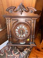 Front view of vintage wooden wind-up wall clock showing carved pediment, columns, and clock face with Roman numerals.