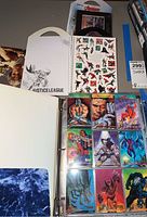 Top view showing binder with X-Men trading cards in protective sleeves, Avengers stickers sheet and Justice League sticker book.