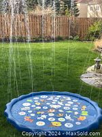 Photo showing blue inflatable circular splash pool with colorful letters and animal graphics inside, spraying multiple water streams upwards in a grassy outdoor yard.