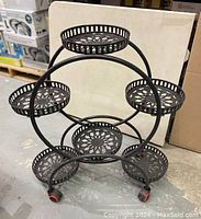 Black metal plant stand showing six tiers and round perforated trays with floral designs. Stand on caster wheels with brake locks.
