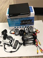 Top view showing DVR unit, 4 cameras, remote control, power adapters, and cables arranged on floor with original box behind