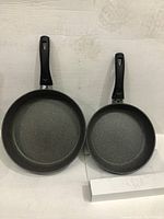 Two frying pans, large and smaller, with black non-stick coating and black handles placed on a white background.