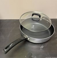Aluminum sauté pan with non-stick black interior, black riveted handle and glass lid with black knob against a neutral background.