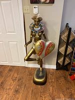 Full view of 52-inch tall bronze-colored African statue showing figure kneeling on one leg, holding spear and shield, with large circular beaded neck piece and head dress.