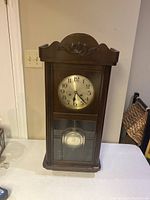 Full view of the antique wooden wall clock showing top carved crest, clock face, and pendulum behind glass door.