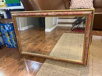 Framed rectangular mirror with patterned reddish-brown and gold decorative frame, positioned on the floor showing reflection of room.