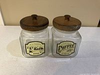 Photo of two square glass containers with wooden lids, labeled for tea and coffee, on a white surface.