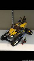 Front angle of the Lego vehicle showing tracked wheels, yellow and brown components, and minifigure with accessory weapon