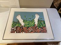 Framed silkscreen print showing two feet with red toenails amid green leaves and crushed Coca-Cola cans under glass in white frame.