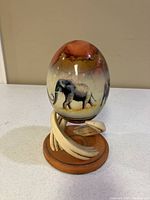 Front view of painted ostrich egg depicting elephants, mounted on resin 'polystone' stand with curved tusk-like elements on wooden base.