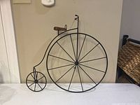 Full view of the penny farthing wire wall display leaning against a wall, showing its large front wheel and smaller rear wheel with wooden seat.