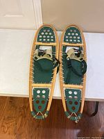 Pair of wooden frame snowshoes with green webbing and green bindings, laid side by side on a light surface.