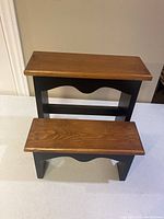 Front view of two-step wooden stool showing natural wood top steps and black painted sides with decorative cut-out details.