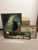 Three framed art pieces leaning against wall: sailboat painting, framed map, and long decorative mirror with coastal painting.