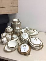 Full view of stacked dinnerware and serving pieces on table, showing multiple plates, cups, and serving pieces with gold and floral trim.