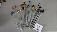 Full set of 28 cocktail stir sticks arranged side by side showing variety of colorful glass and metal decorative toppers.