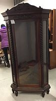 Front view of dark wooden corner cabinet with glass door, decorative carved top, twisted vertical trim columns, and four legs.