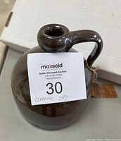 Full view of the antique brown ceramic crock jug with handle and round body on a grey surface with a white box background. Shows attached tag with string.