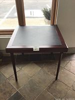 Full view of square folding table near window, showing black leatherette top framed by reddish-brown polished wood and straight folding legs.