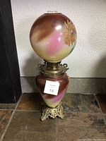 Full view of the antique oil lamp showing pink and brown gradient glass globe and base with floral painting, brass-toned ornate metal base.
