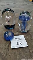 Three paperweights displayed on floor: two clear glass with internal pink flower and blue jellyfish designs, one blue plastic with dandelion seed inside.