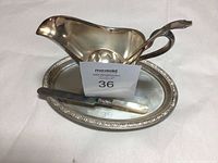 Silver gravy boat on oval platter with mother of pearl handled spoon resting on platter, displayed on a white surface.