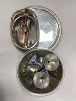 Overview of set including oval serving dish with silverware inside, round serving platter underneath, and four silver candlestick holders in a round container.