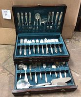 Wooden chest open showing upper and lower velvet-lined compartments filled with flatware pieces