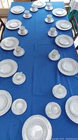 Full dining table arrangement showing 12 sets of cups, saucers, bread plates, and dinner plates on a deep blue tablecloth.