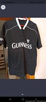 Front view of black Guinness shirt with white Guinness text and harp logo, hung on hanger.