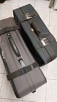 Two large traditional suitcases in black and dark gray with buckle straps and handles placed on floor